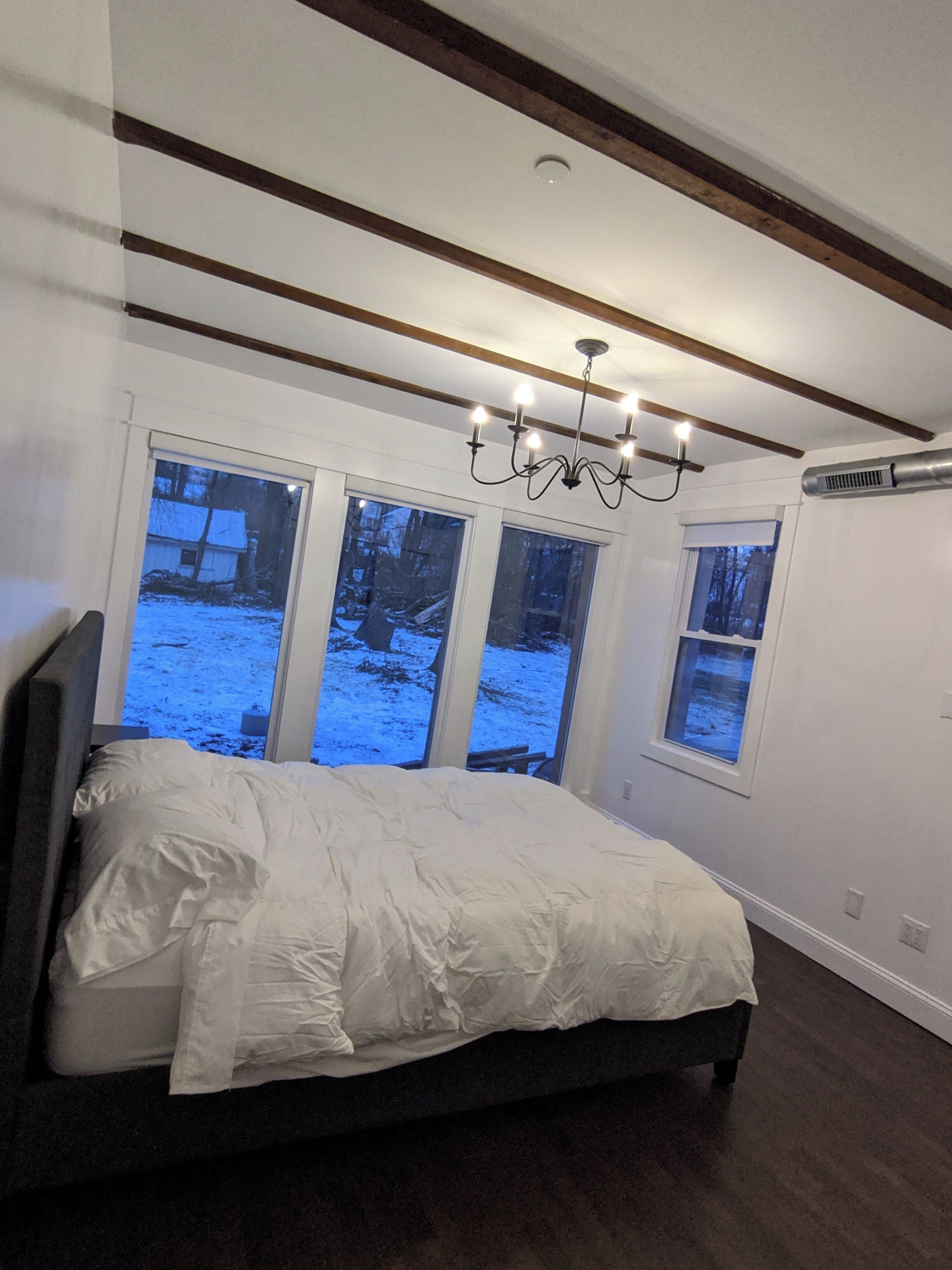 A bedroom with a bed, a chandelier, large windows, and a ceiling vent.