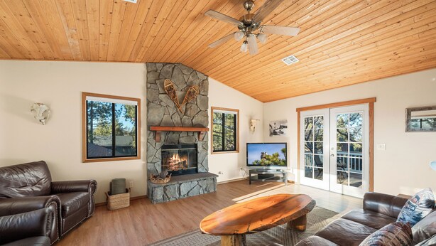 A spacious living room with a stone fireplace, leather sofas, a wooden coffee table, and large windows offering a view of the outdoors. - Beau Bear (Big Bear City)