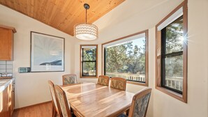 A dining area with a wooden table and chairs, large windows with a view of trees, and a ceiling with wooden beams. - Beau Bear (Big Bear City)