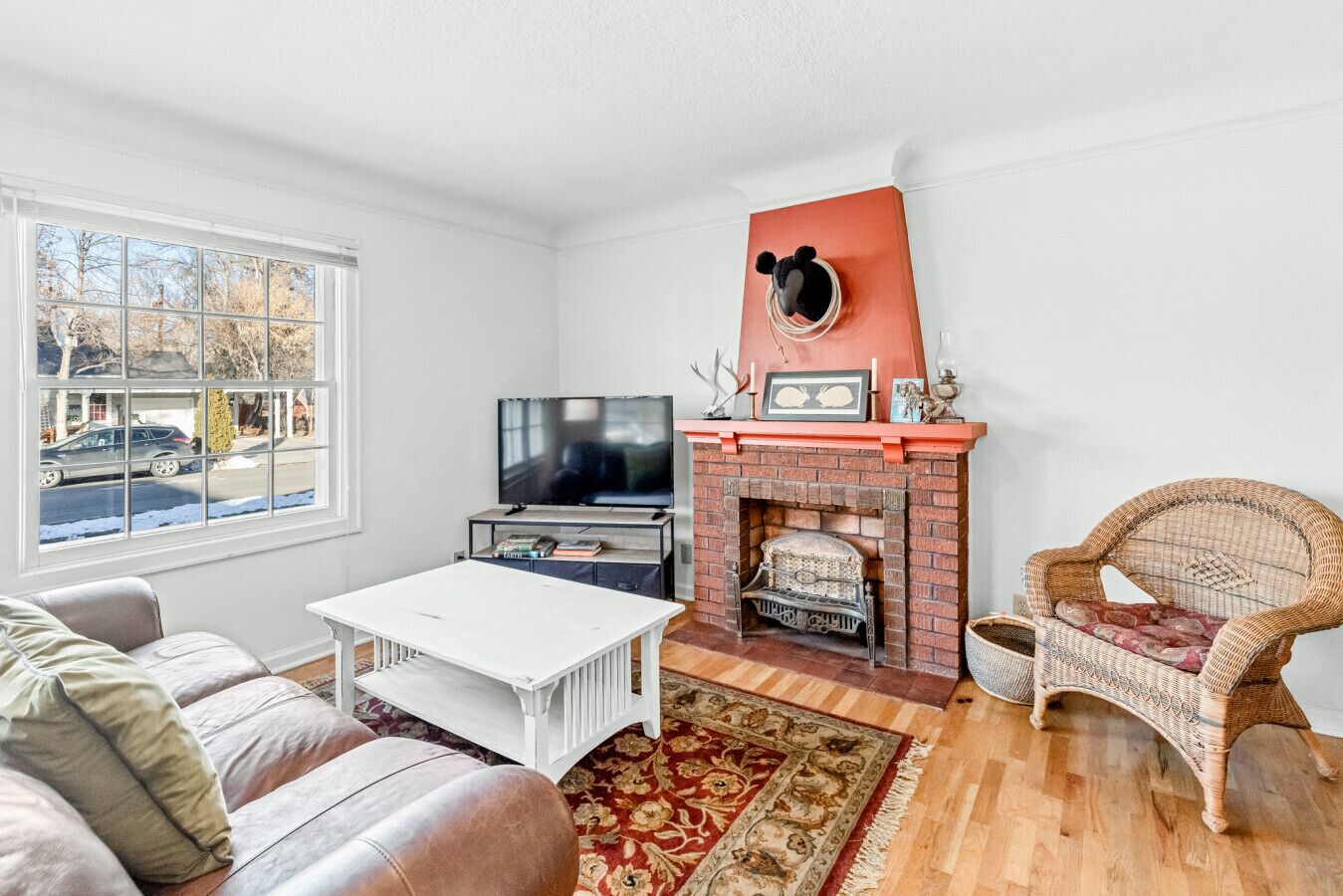 Ein Wohnzimmer mit Kamin, Fernseher, weißem Couchtisch, Korbstuhl und Blick auf die Straße durch ein großes Fenster.