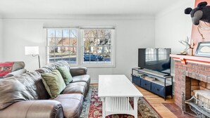 Een woonkamer met een leren bank, een witte salontafel, een tv op een meubelstuk, een open haard en uitzicht op de straat door grote ramen.