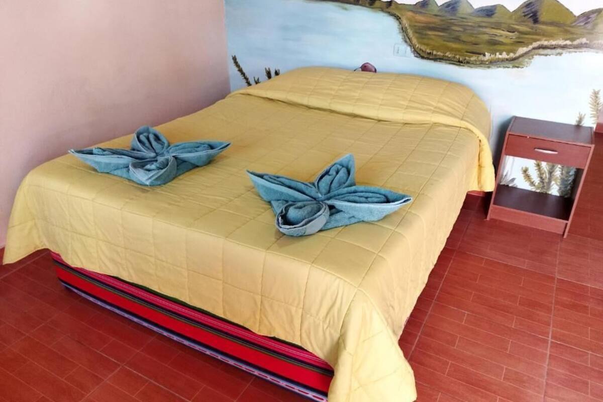 Una cama con una colcha amarilla y dos toallas azules con forma de hojas, una falda a rayas rojas y negras, un cuadro de un paisaje en la pared y una mesita de noche de madera con un cajón.