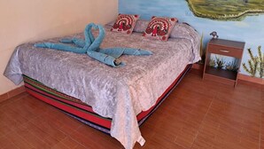 A bed with a patterned bedspread and pillows, a small bedside table with a plant, and a mural of a landscape on the wall. - Hostal Amigo Lucho  (San Pedro de Atacama)