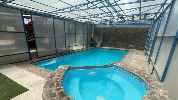 An indoor swimming pool with a stone hot tub, surrounded by glass walls and a metal roof. - Hostal Amigo Lucho  (San Pedro de Atacama)