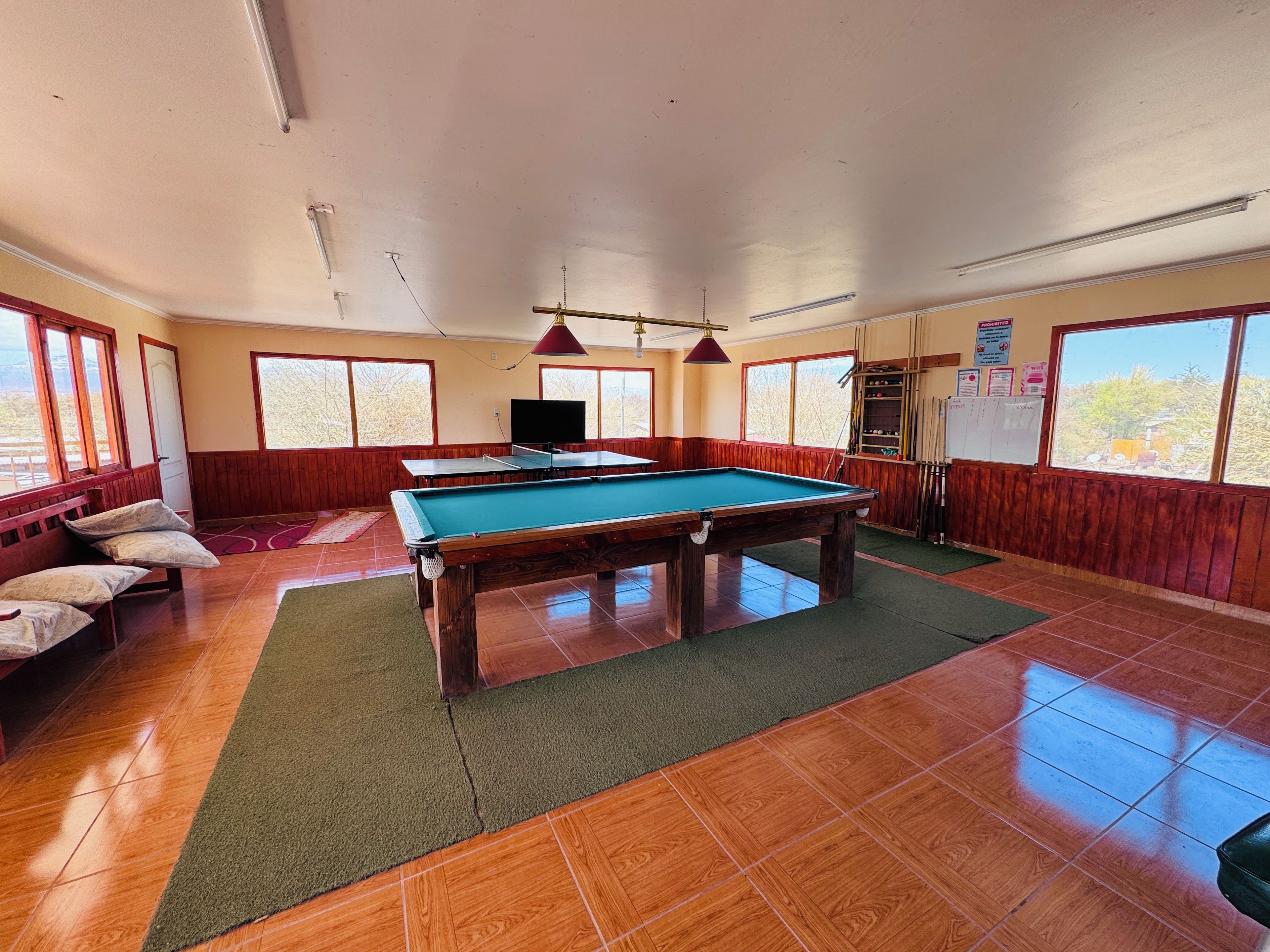 A pool table in a room with wooden flooring, a green carpet, and a television.
