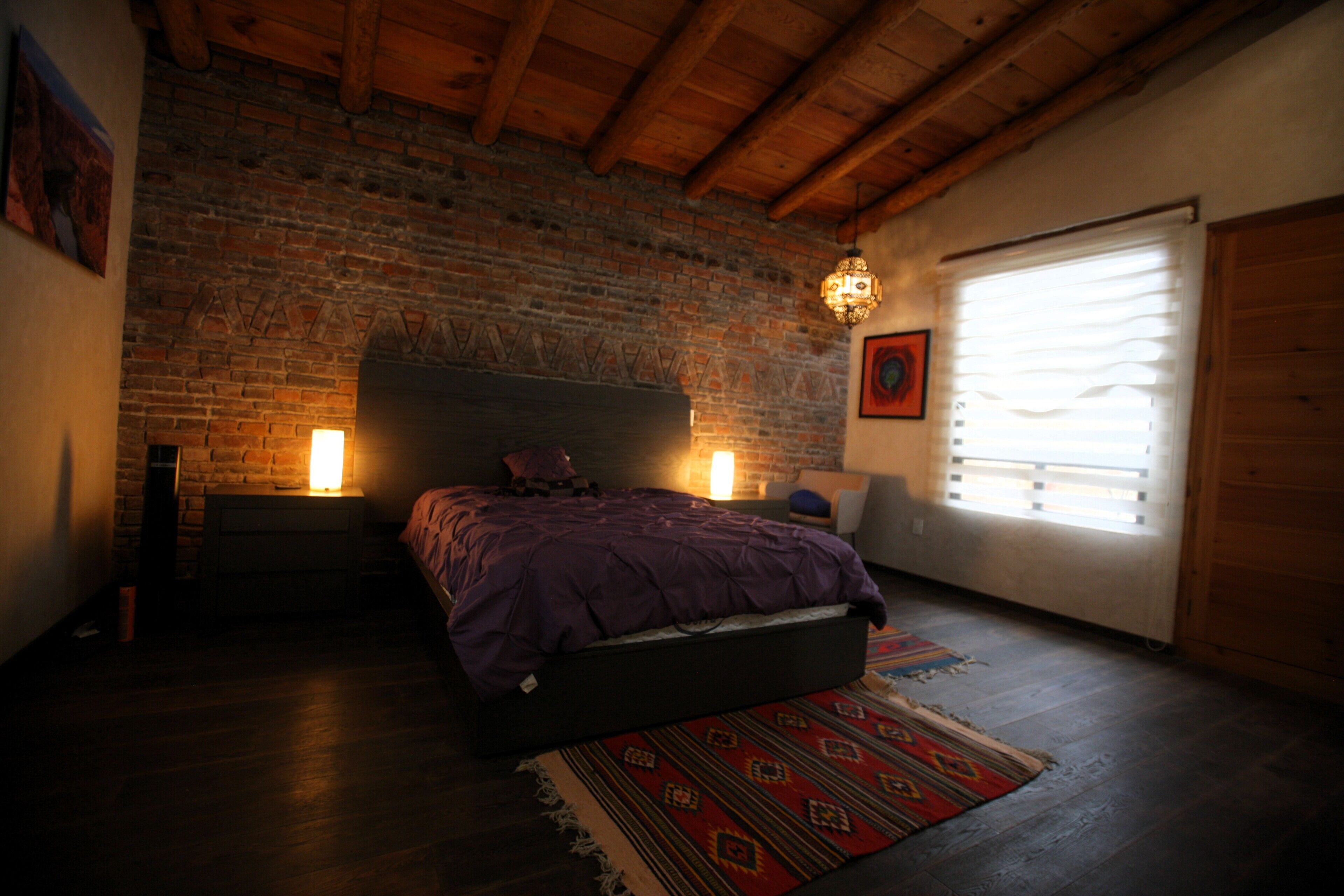 Un dormitorio con pared de ladrillo, vigas de madera, cama con colcha morada, alfombra y ventana con persianas.