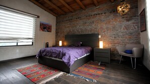 A bedroom with a bed, nightstands, a chair, a patterned rug, a window with blinds, and a brick wall. - Maceiras House (San Cristóbal de las Casas)