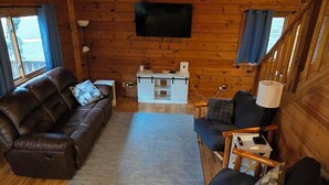 A cozy cabin interior with a wooden floor, a sofa, a chair, a small table, and a TV mounted on the wall.