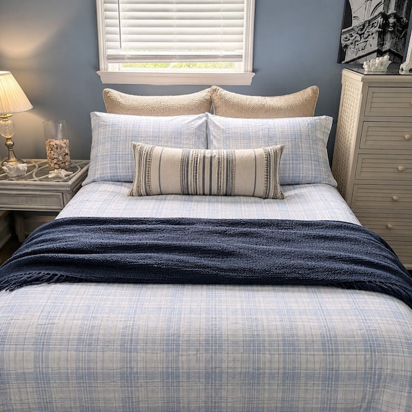 Una cama con colcha a cuadros azul y blanco, dos almohadas, una manta azul oscuro, una mesita de noche con lámpara y un aparador con cajones.