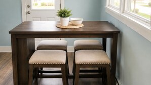A dining area with a wooden table, two upholstered chairs, a potted plant, a bowl, and a wall clock.