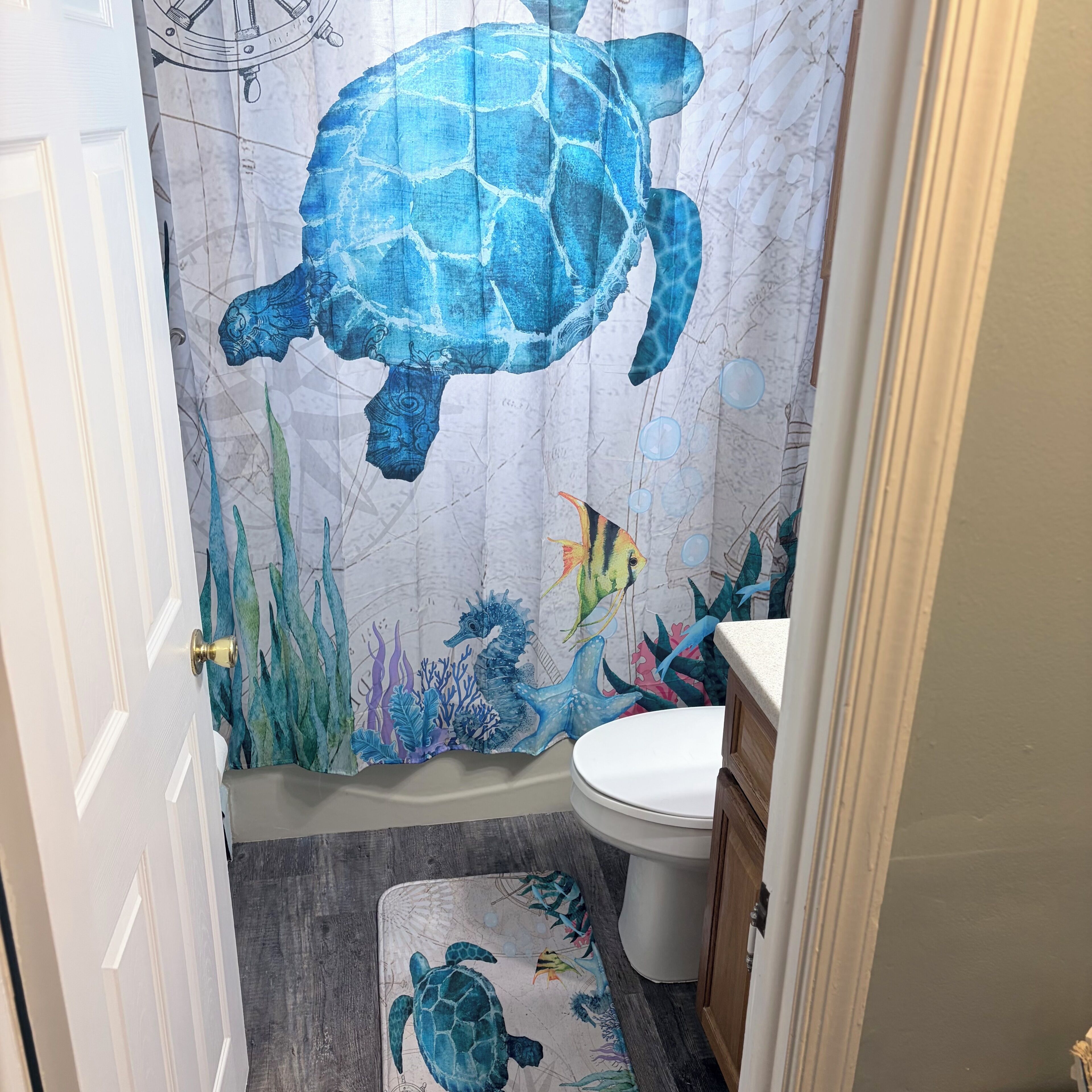 A bathroom with a seahorse and turtle shower curtain, a wooden floor, and a white toilet.