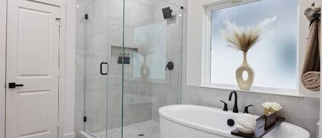 A modern bathroom with a glass-enclosed shower, a freestanding bathtub, and a window with a vase of flowers.