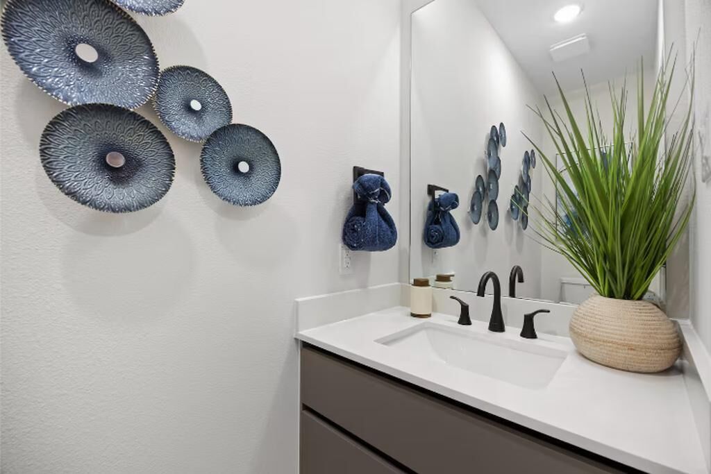 Un bagno con un lavandino bianco, rubinetti neri e un grande specchio. Sul mobile ci sono asciugamani decorativi blu e una pianta in vaso.