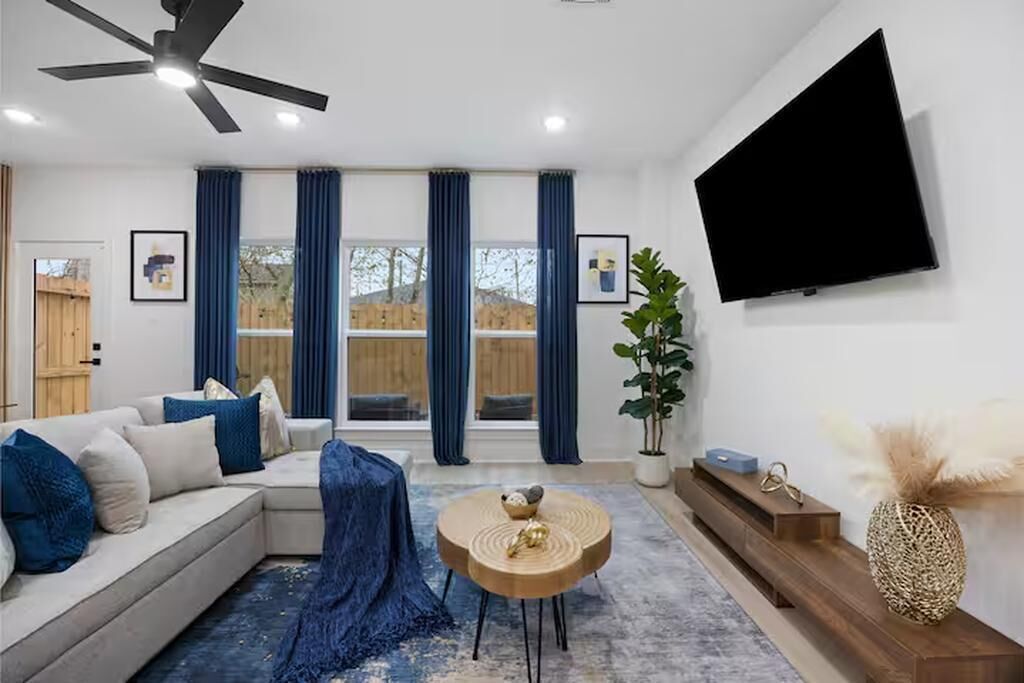 Ein modernes Wohnzimmer mit einem Ecksofa, einem runden Couchtisch, einem Deckenventilator, einem Flachbildfernseher und großen Fenstern mit blauen Vorhängen.