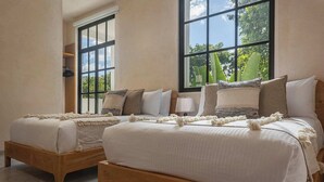 Dos camas de madera con sábanas blancas y flecos, una ventana con vistas a la vegetación y una lámpara fijada a la pared.