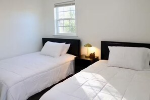 Twee eenpersoonsbedden met witte lakens, een nachtkastje met een lamp en een raam met natuurlijk licht.