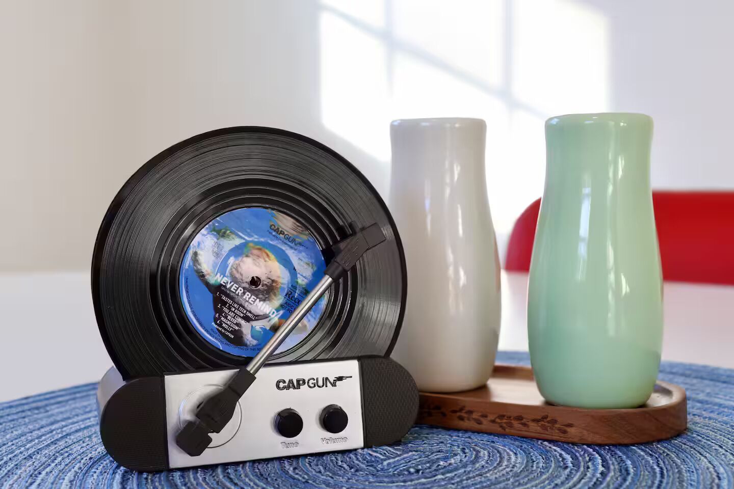Une platine vinyle avec un disque en vinyle, un vase blanc et un vase vert, le tout posé sur une surface bleue à motifs.