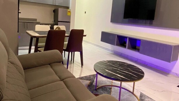 A living room with a sofa, a dining table with chairs, a TV mounted on the wall, and a ceiling fan. - One Bed room Apartment, BH Apartments (Dansoman)