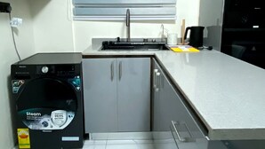 A compact kitchen with a built-in sink, grey cabinets, a black appliance, and a window with blinds. - One Bed room Apartment, BH Apartments (Dansoman)