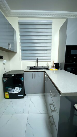 Cocina compacta con lavaplatos integrado, gabinetes grises, electrodomésticos negros y una ventana con persianas.