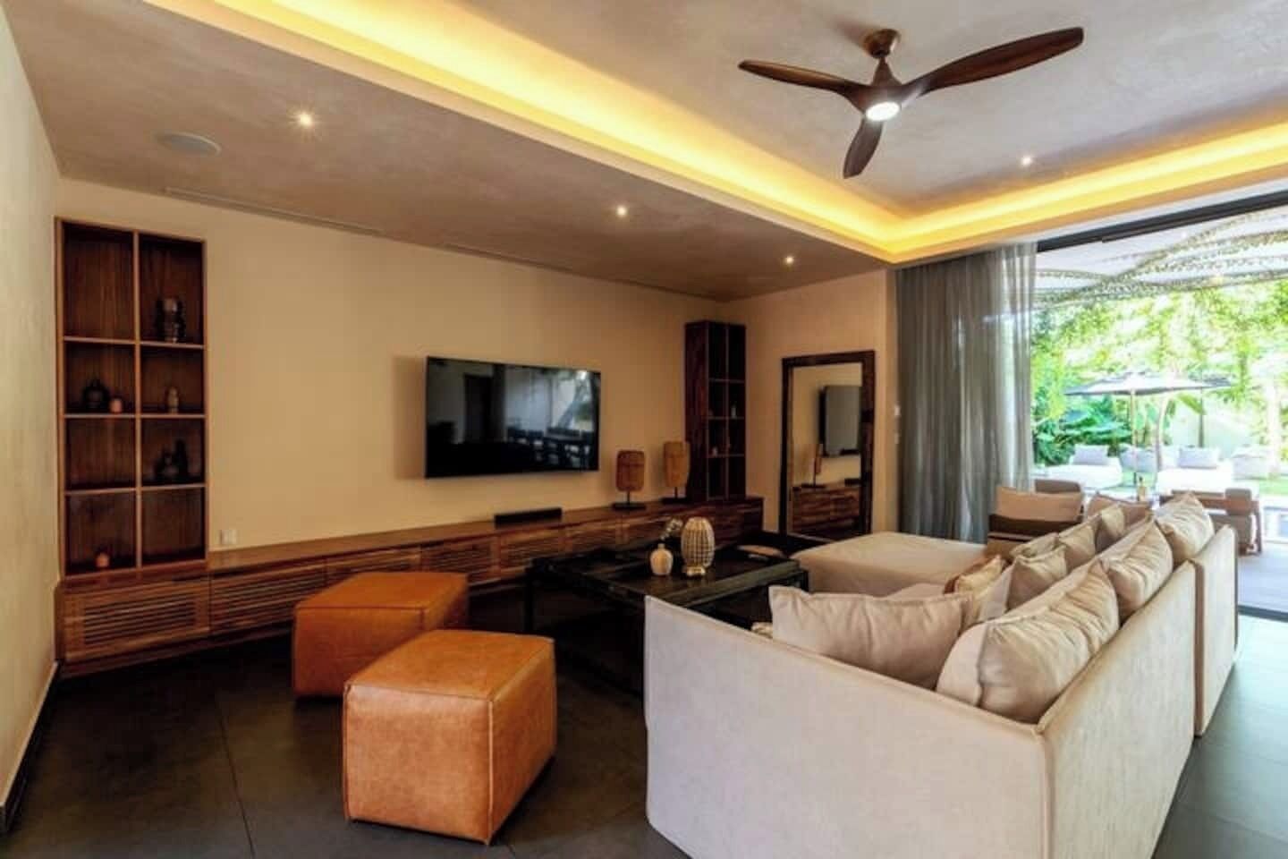 A modern living room with a sofa set, a television, a ceiling fan, and a large window with a view of greenery.