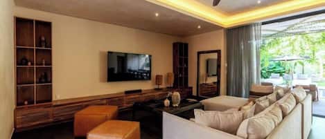 A modern living room with a sofa set, a television, a ceiling fan, and a large window with a view of greenery.