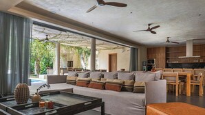 Een ruime woonkamer met een bank, salontafel en stoelen, een keuken met houten kastjes, en uitzicht op groen.