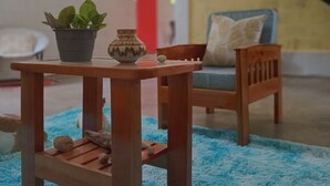 Une petite table en bois, sur laquelle se trouvent un pot de fleurs et un bol décoratif. Un tapis bleu recouvre le sol, une chaise en bois est placée à proximité, et un escalier se trouve au fond de la pièce.