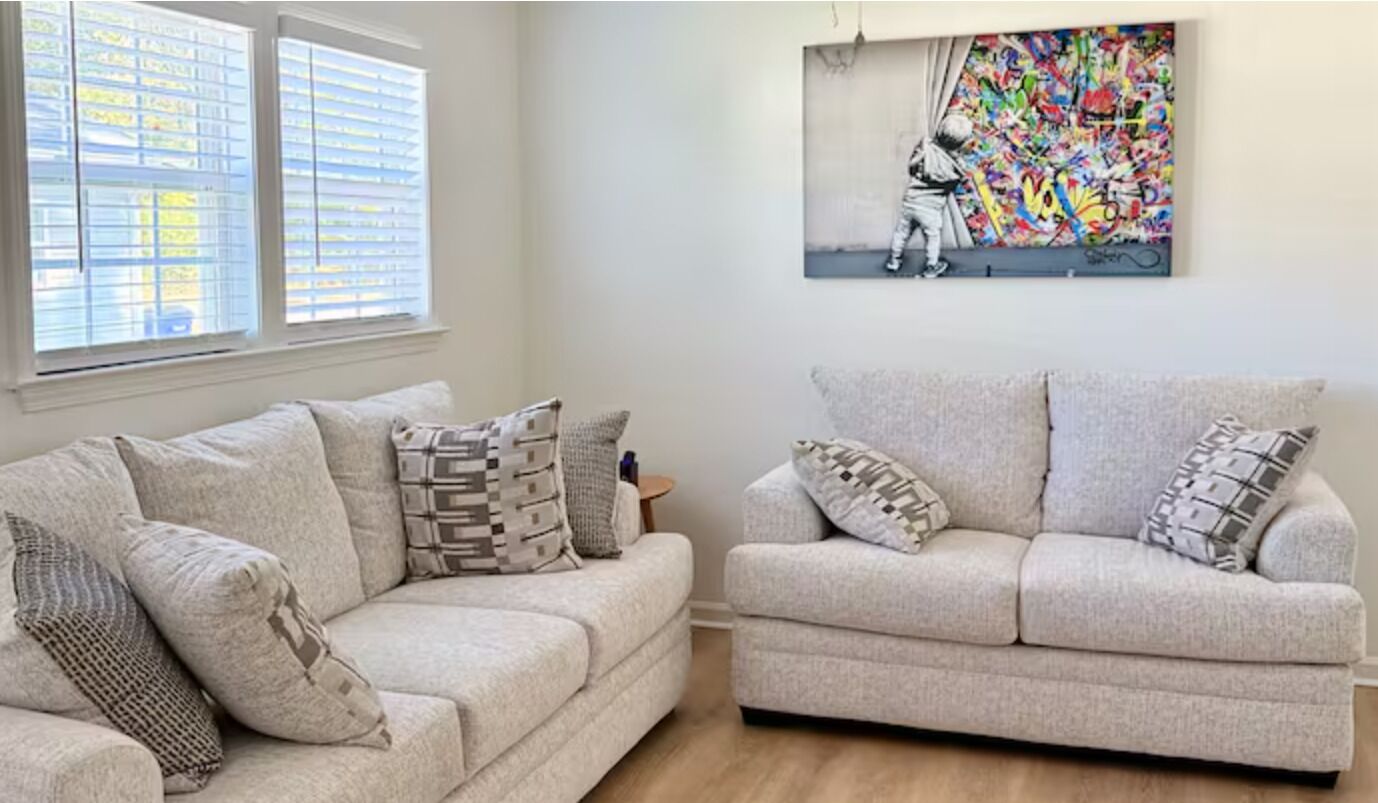 A living room with two light-colored sofas, patterned cushions, a colorful abstract painting on the wall, and a window with blinds.