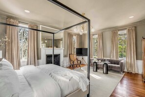 Ein Schlafzimmer mit Himmelbett, Kamin, Fernseher, Sofa und einem großen Fenster mit Vorhängen.