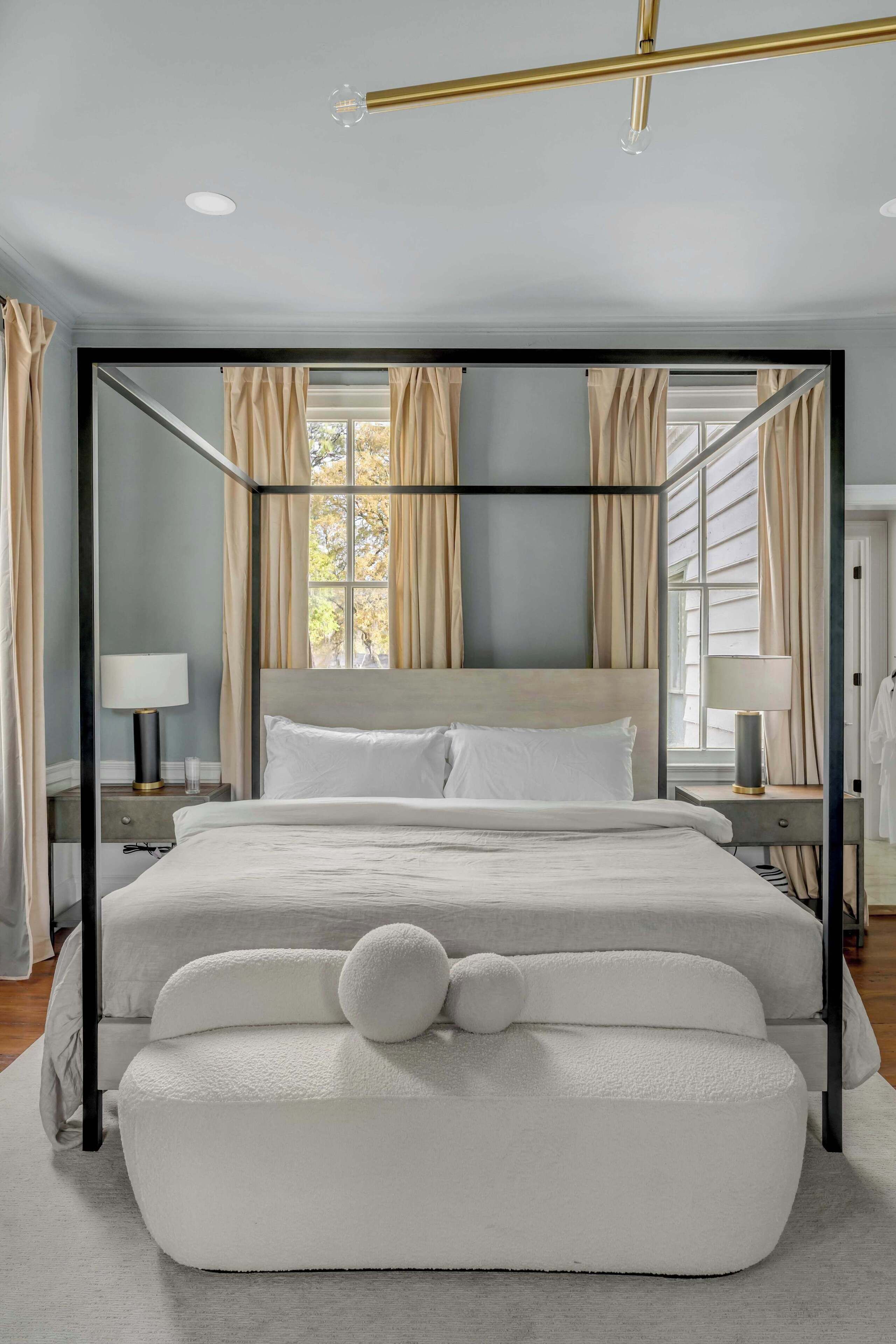 Un lit à baldaquin avec une tête de lit et un pied de lit blancs, une table de chevet avec une lampe, et une vue sur l’extérieur par une fenêtre ornée de rideaux.