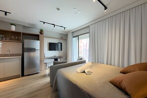 A modern hotel room with a bed, a desk, a chair, a TV, and a kitchen area. - Ape em Perdizes 8 andar prox. ao Allianz (São Paulo)