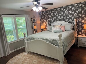 Een slaapkamer met een bed, een plafondventilator, een nachtkastje, een lamp en een raam met gordijnen.