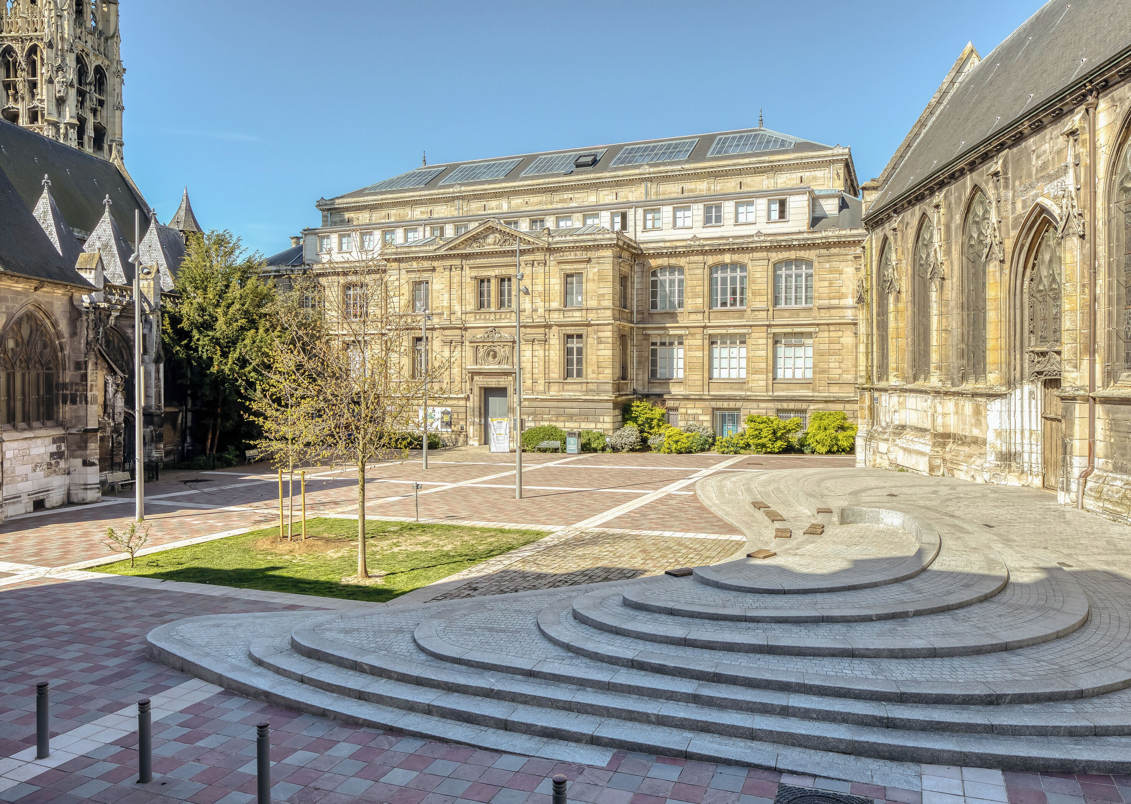 A courtyard with a central circular stone feature, a building with classical architecture, and a tree.