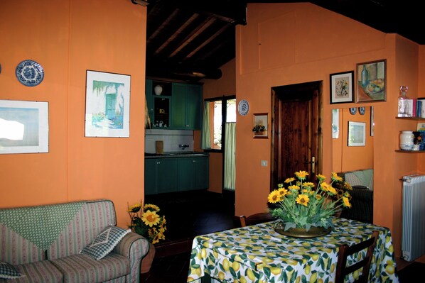 A cozy living room with a sofa, a dining table with a floral tablecloth, and a kitchen in the background.