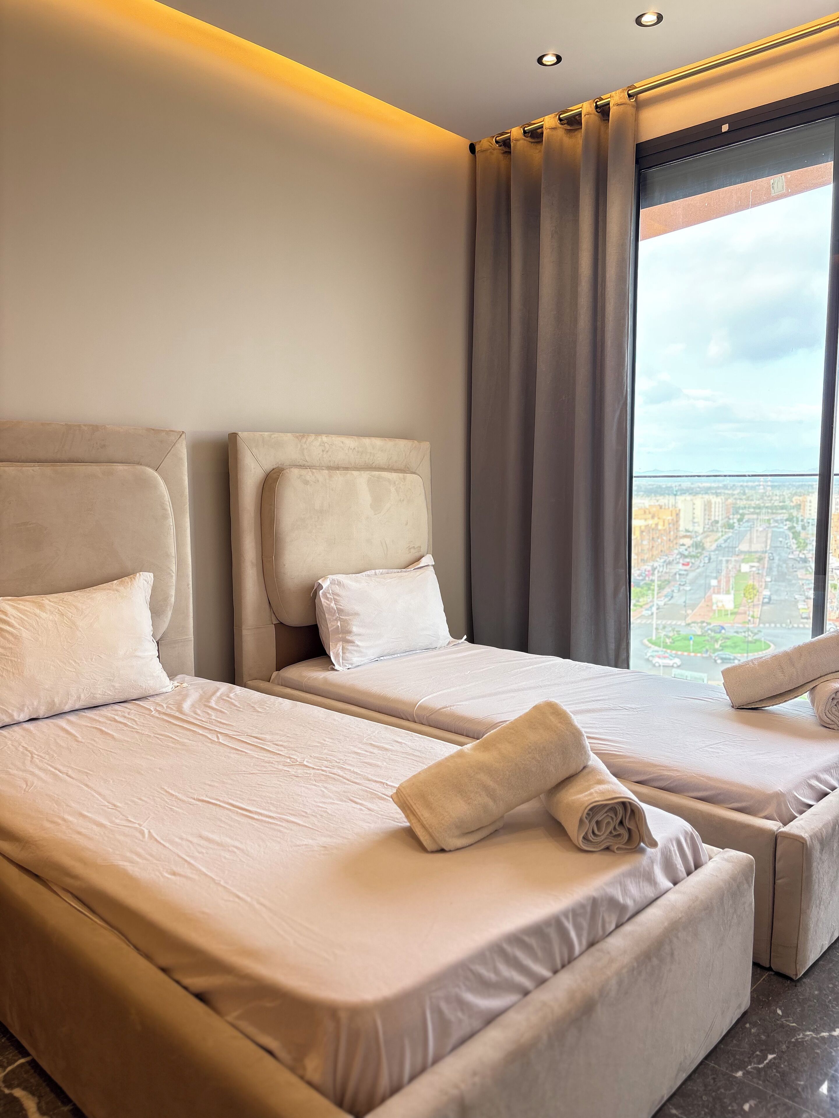 Quarto de hotel com duas camas, cada uma com cabeceira, uma janela grande com cortinas e vista para a paisagem urbana.