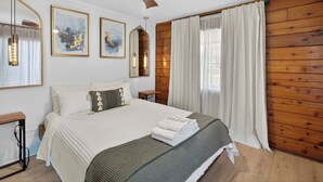 Une chambre avec un plancher en bois, un lit avec une literie blanche, un ventilateur de plafond et deux appliques murales.