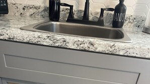 A kitchen with a granite countertop, a stainless steel sink, and a storage cabinet containing a white laundry basket, a yellow bottle, a purple bottle, and a box. - Cozy apartment in Valrico (Valrico)