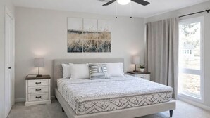 A bedroom with a bed, two bedside tables, a ceiling fan, a window with curtains, and a wall-mounted artwork. - Guesthouse – 5BR Lower Level (2BR+3BR) (Overland Park)