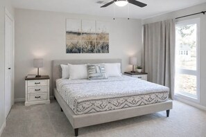 Une chambre à coucher comprenant un lit, deux tables de chevet, un ventilateur de plafond, une fenêtre avec des rideaux et une œuvre d’art accrochée au mur.
