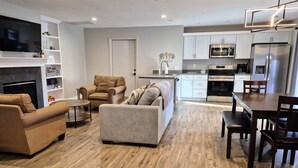 A modern living room with a fireplace, a sofa, a coffee table, and a dining area with a table and chairs. - Guesthouse – 5BR Lower Level (2BR+3BR) (Overland Park)