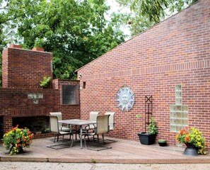Une terrasse en briques avec une table et des chaises, un décor mural représentant un soleil et des plantes en pot.