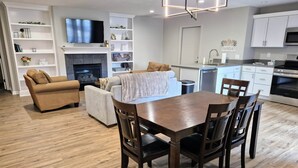 A modern living room with a fireplace, a dining table with chairs, a sofa, and a TV mounted on the wall. - Guesthouse – 5BR Lower Level (2BR+3BR) (Overland Park)