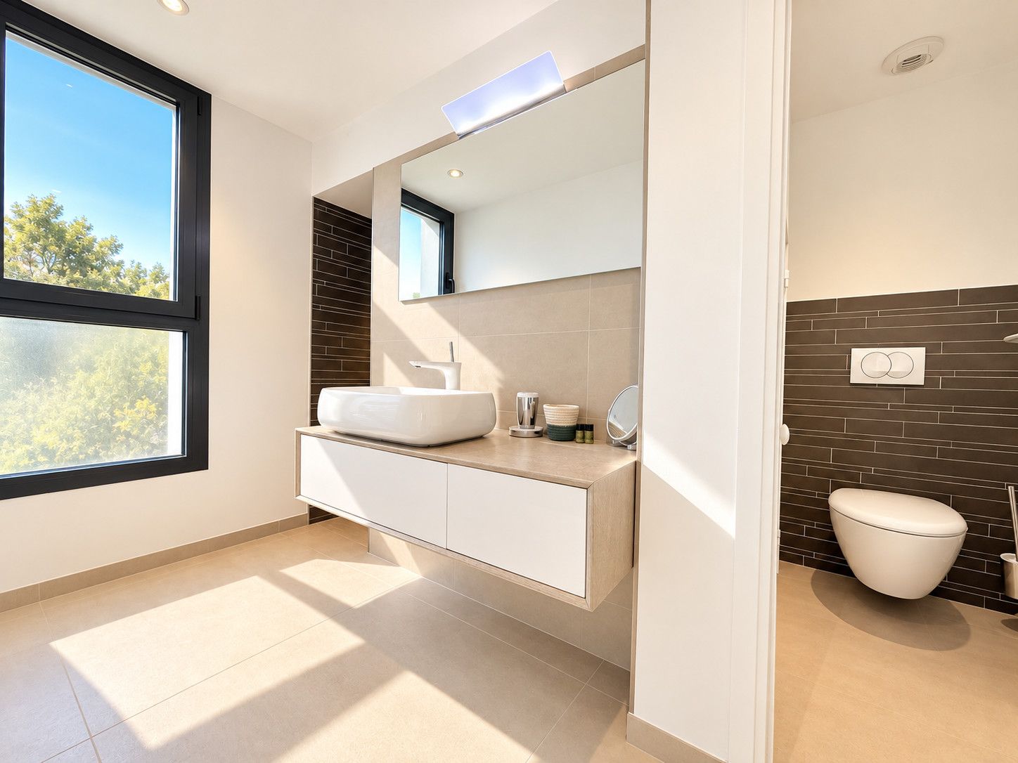 A modern bathroom with a large window, a white sink, a mirror, and a toilet.