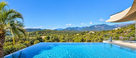 A clear blue infinity pool with a wooden deck, surrounded by lush greenery and a mountainous backdrop.