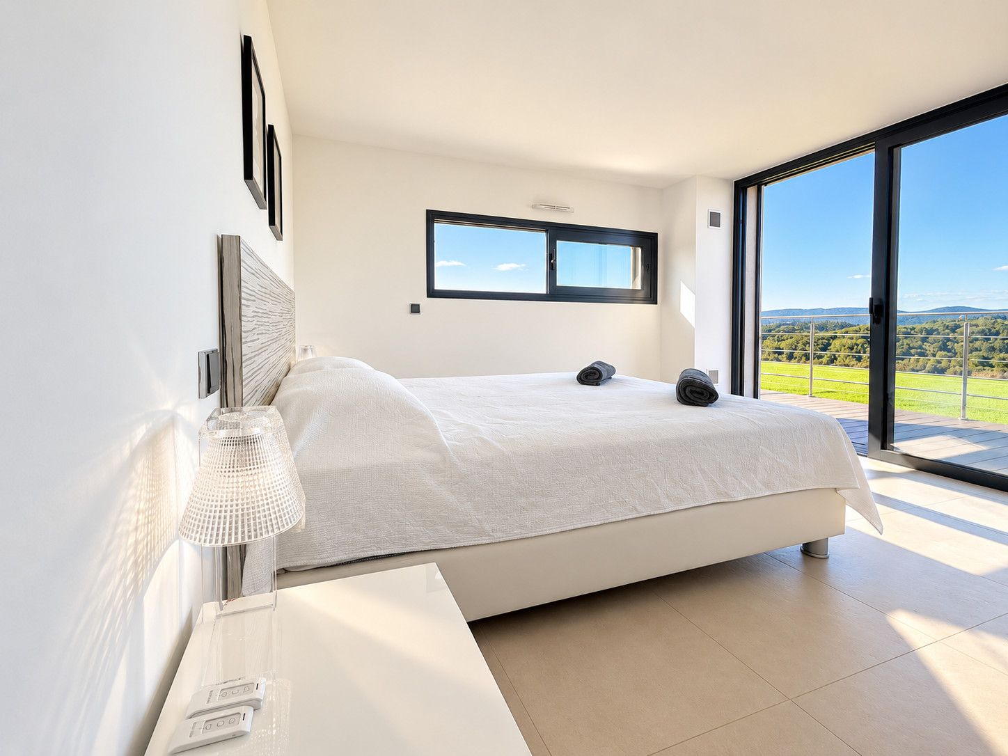 Une chambre moderne avec un grand lit, une table de chevet avec une lampe, et une vue sur un paysage verdoyant par une grande fenêtre.