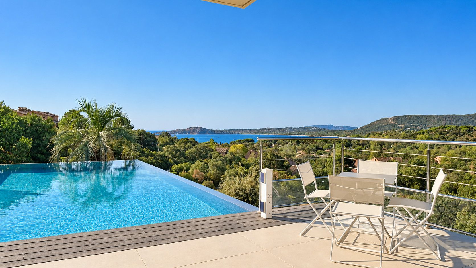 An infinity pool with a clear view of the sea and hills, a wooden deck, and a table with chairs.