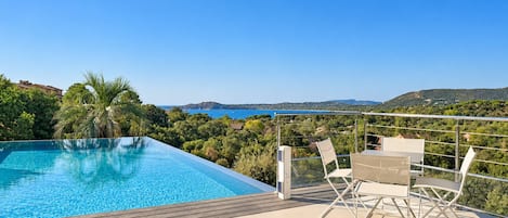 Ein Pool ohne Ende mit freiem Blick auf Meer und Hügel, eine Holzterrasse und ein Tisch mit Stühlen.