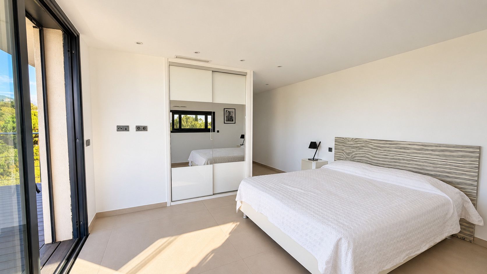 Ein Schlafzimmer mit einem Bett, einem Nachttisch, einem Spiegel und einer Schiebetür, die auf einen Balkon führt.