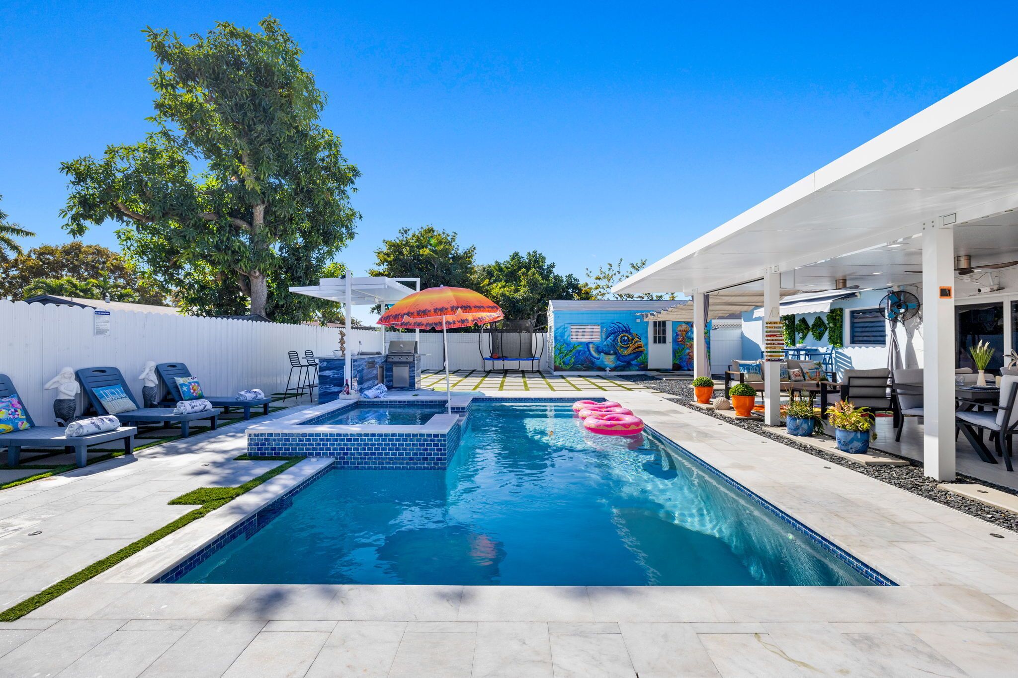 A swimming pool with lounge chairs and umbrellas, surrounded by a white fence and potted plants.